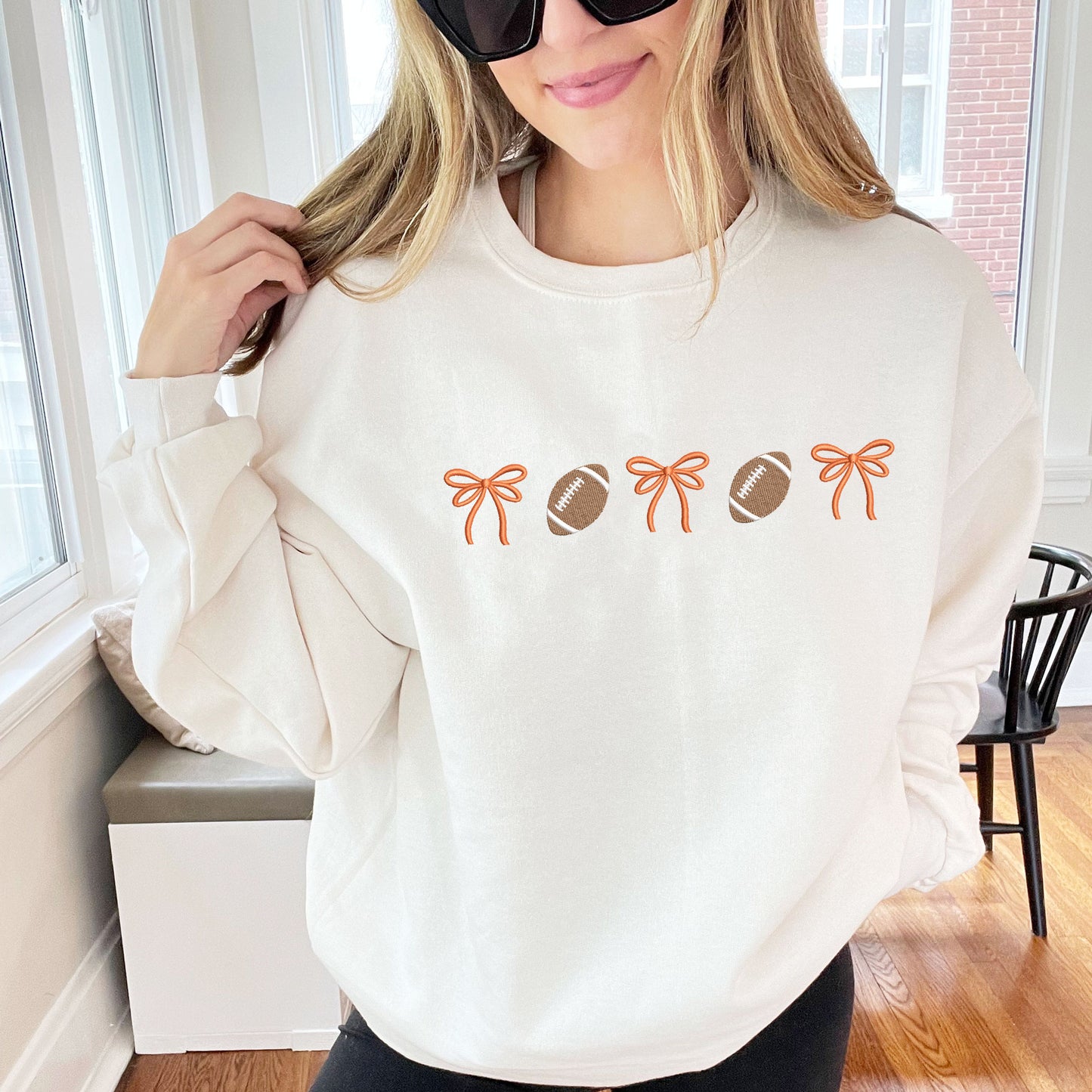 woman wearing a sweet cream crewneck sweatshirt with embroidered alternating bows and footballs design across the chest. The bow are embroidered in orange thread.