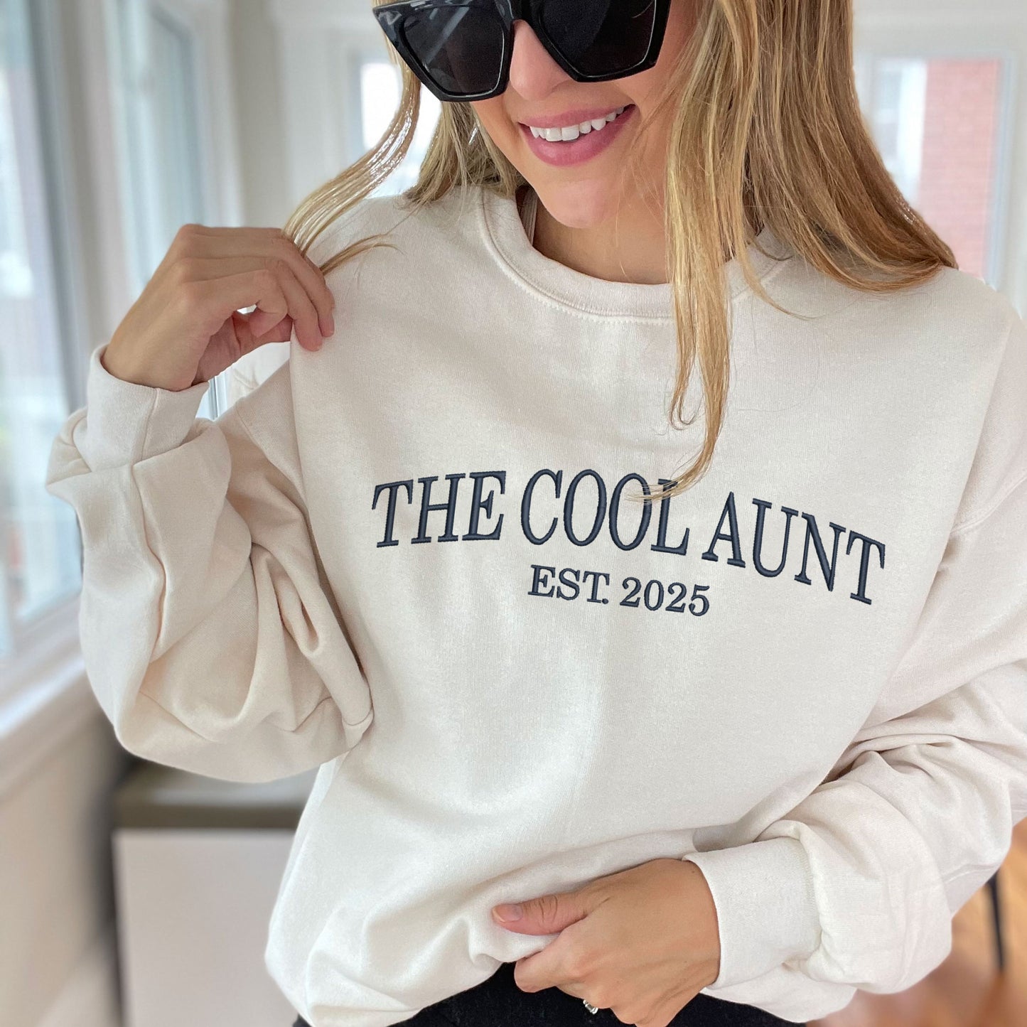 young woman wearing sunglasses and a sweet cream crewneck sweatshirt with embroidered The Cool Aunt Est. 2025 in navy thread large across the chest