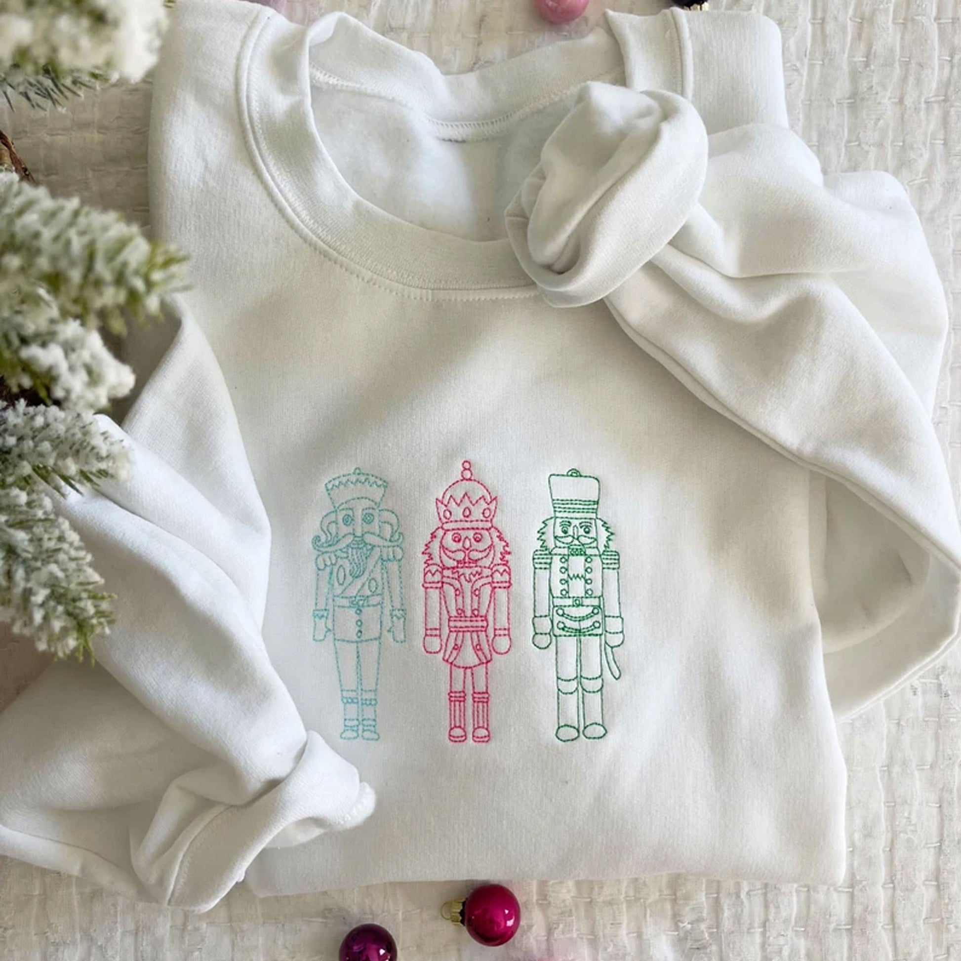 Flat lay image of a white crewneck sweatshirt showing 3 embroidered stitched nutcrackers across the chest in blue, pink, and green thread.