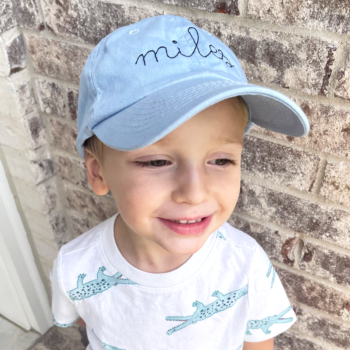 little boy wearing a baby blue youth baseball cap with embroidered name in a stitched cursive font and navy thread