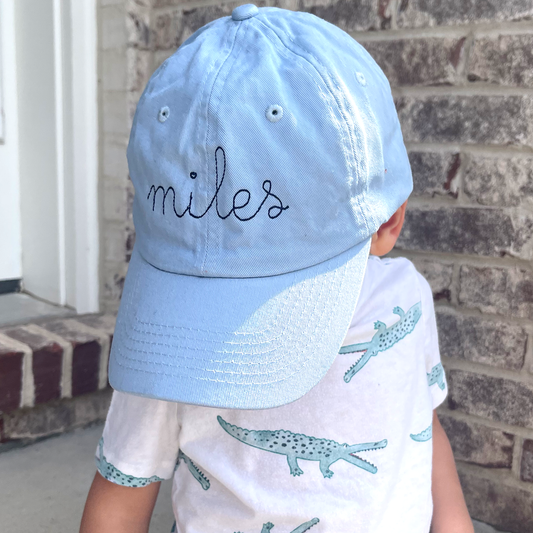 little boy wearing a baby blue youth baseball cap with embroidered name in a stitched cursive font and navy thread 