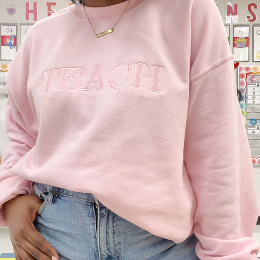 light pink crewneck sweatshirt with embroidered all caps teach in  baby pink tone on tone thread