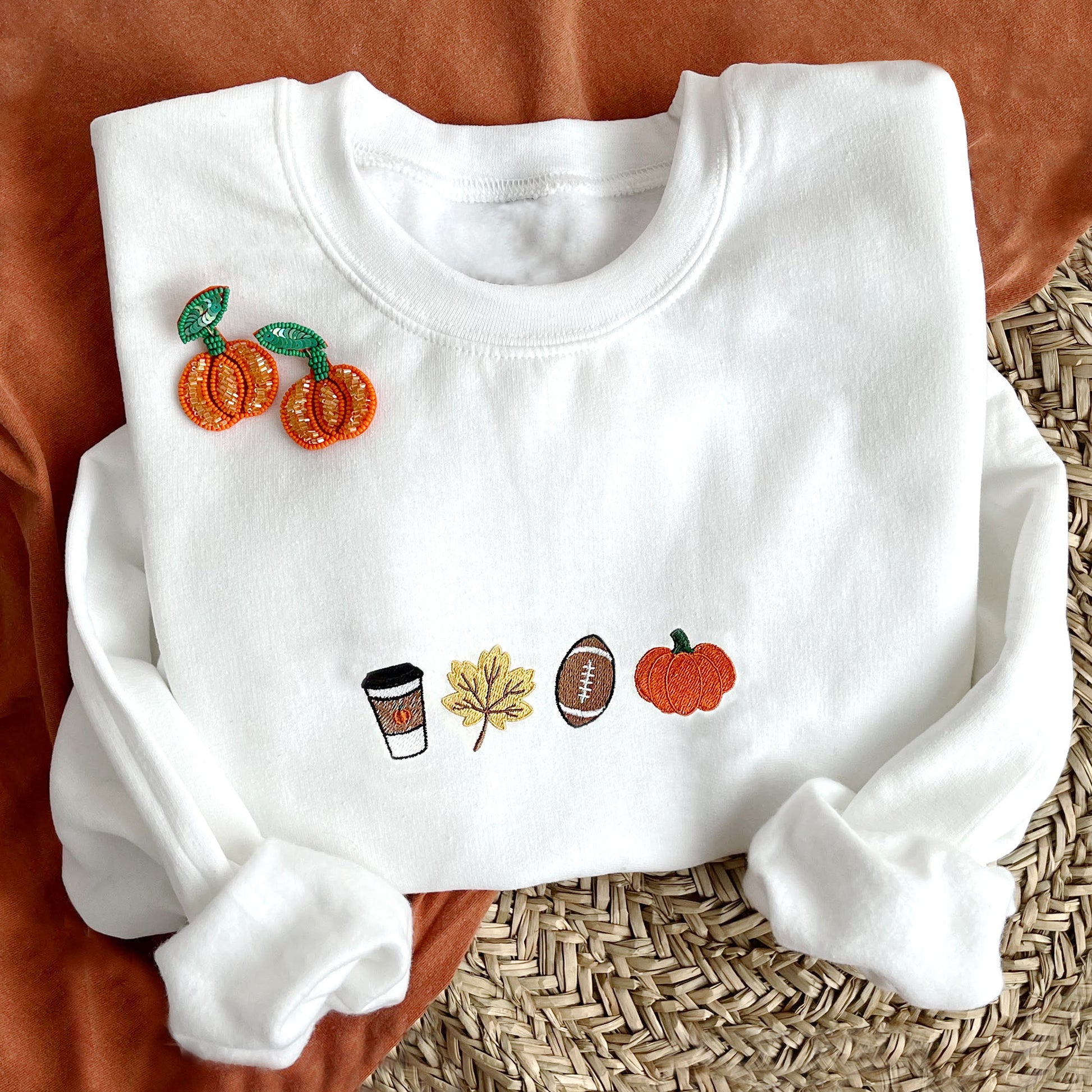 styled flat lay photo of a white crewneck sweatshirt with embroidered coffee, leaf, football, and pumpkin design across the chest.