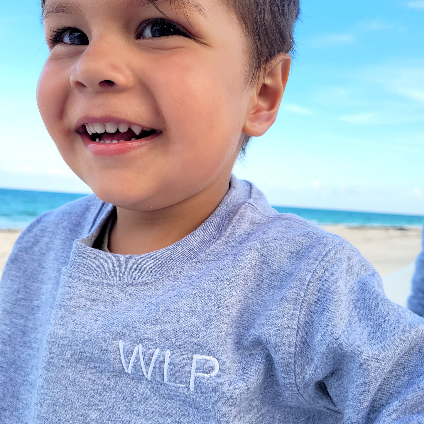 close up of boy smiling wearing light gray crewneck with initials embroidered in arial font
