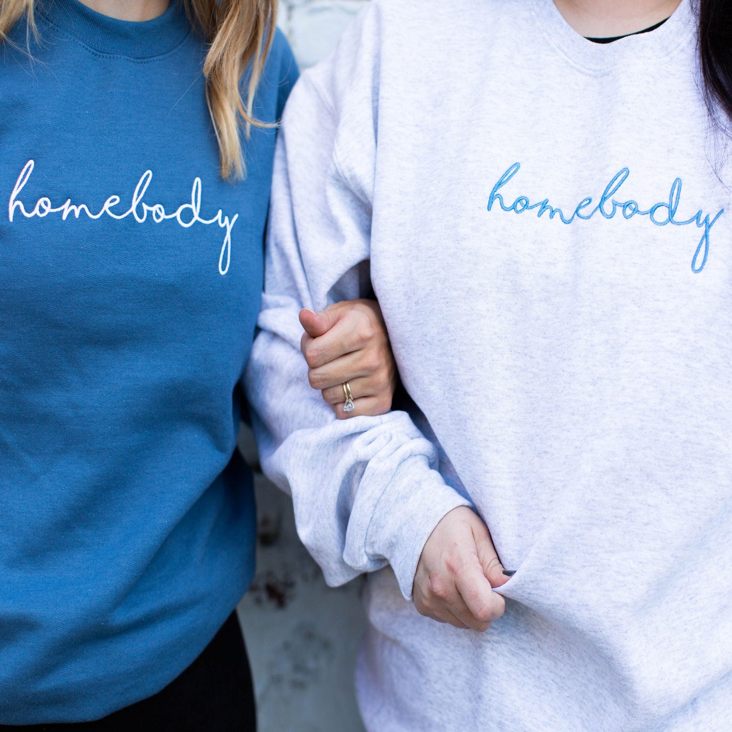 two girls standing side-by-side wearing crewneck pullovers one is blue with homebody embroidered in white thread across the chest and the other is ash gray with homebody embroidered in blue thread across the chest