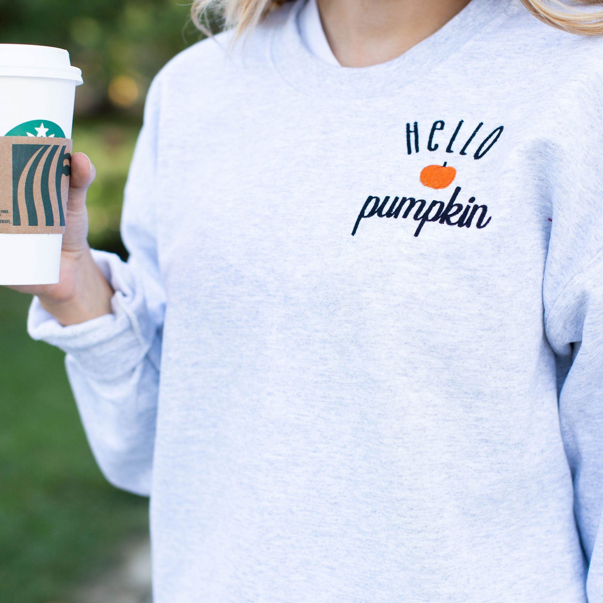 Girl holding travel coffee cup wearing light gray crewneck sweatshirt with hello pumpkin embroidered in black with an orange mini pumpkin between