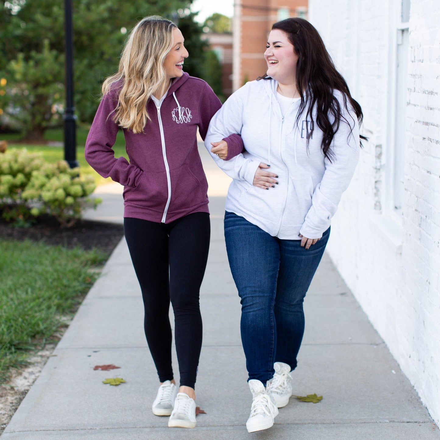 Two girls walking side by side. Blonde wearing a maroon full zip jacket with white monogram embroidery and brunette wearing ash gray full zip jacket with blue monogram embroidery