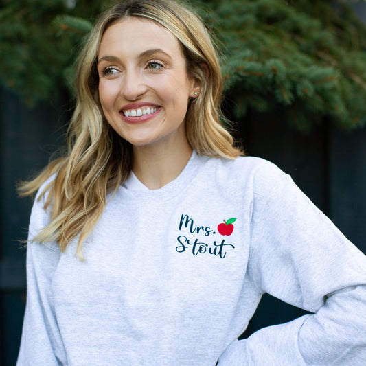 woman wearing a light gray crewneck sweatshirt with teacher name and mini apple embroidery