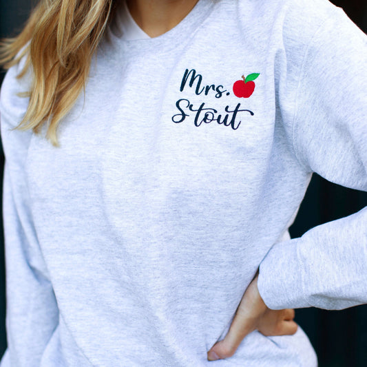 close up of educator's name and small red apple embroidered on a an ash pullover sweatshirt