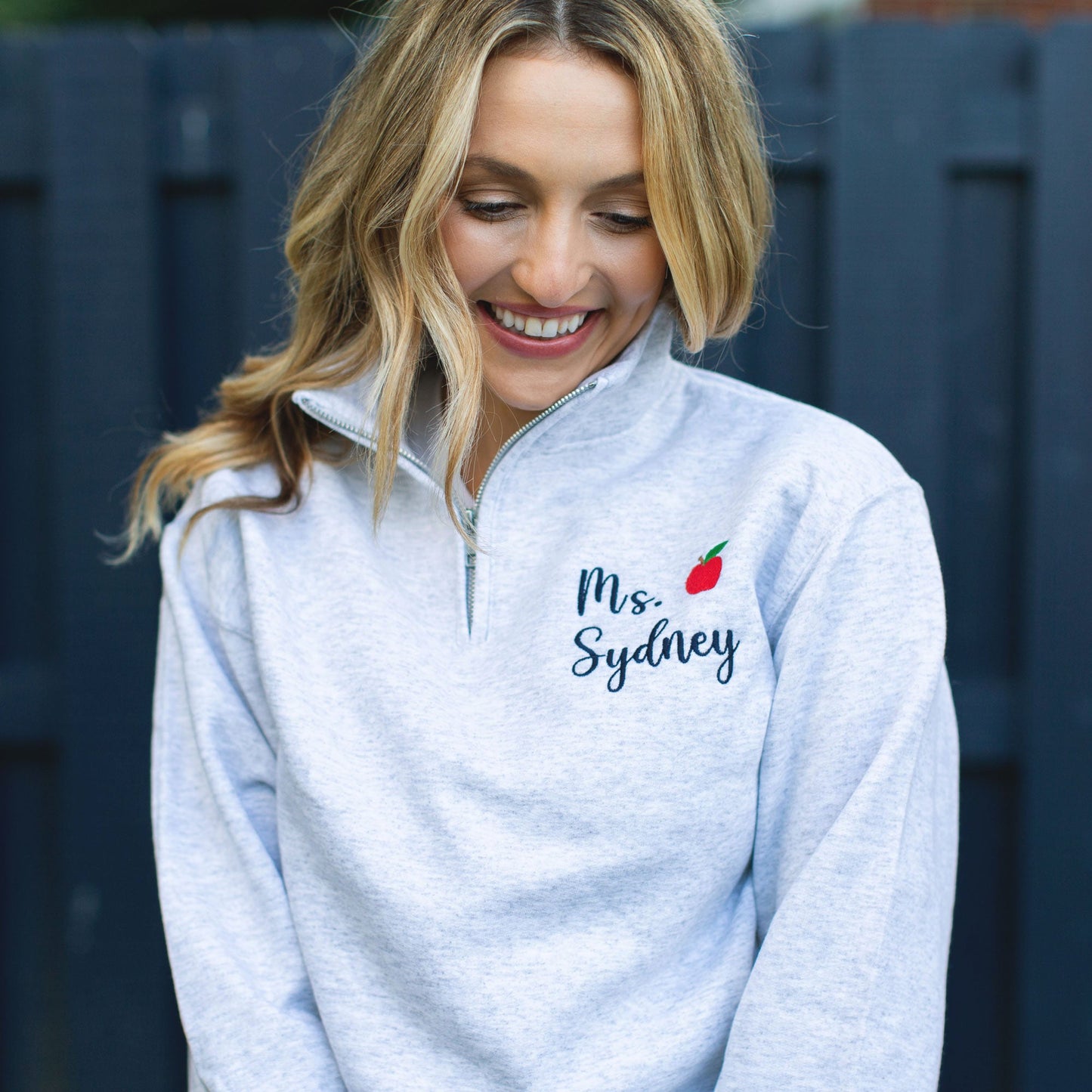 Woman wearing ash gray quarter zip sweatshirt with teacher name and mini apple embroidery