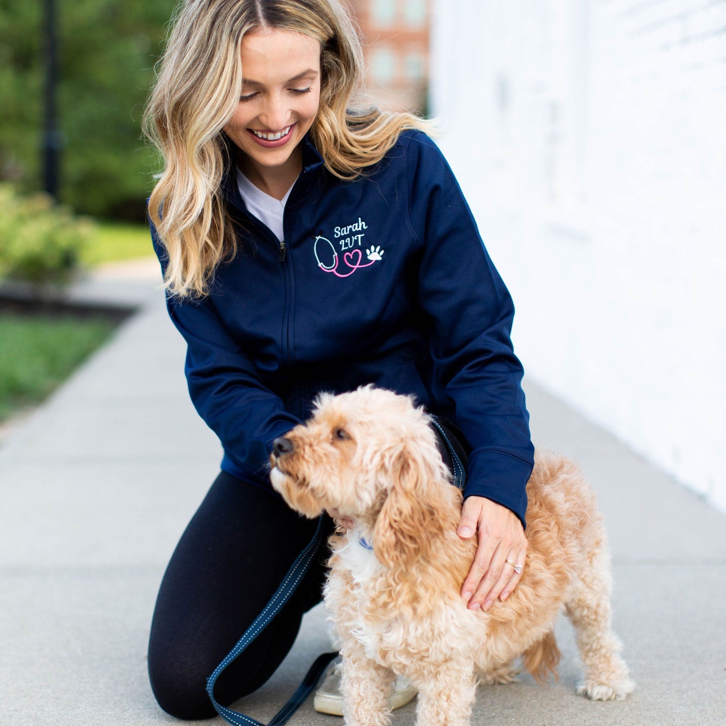 Girl wearing a blue jacket with personalized vet technician name embroidery and paw print stethoscope kneeling next to her small dog