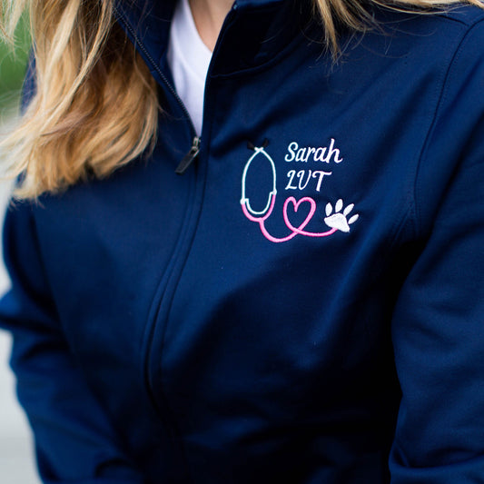 close up of navy full zip polyester fleece jacket showing embroidered bright pink heart stethoscope, white paw, and white name in a script font