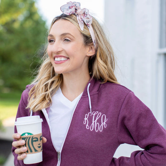 Blonde woman holding a travel coffee cup and wearing a heather maroon full zip jacket with script monogram in white thread