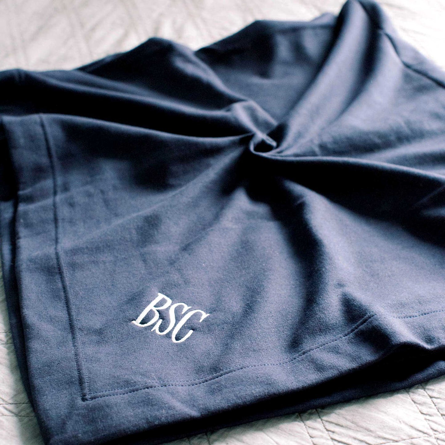 a folded stadium blanket in a dark navy blue on a bed with a custom three letter embroidered monogram in white thread on the corner