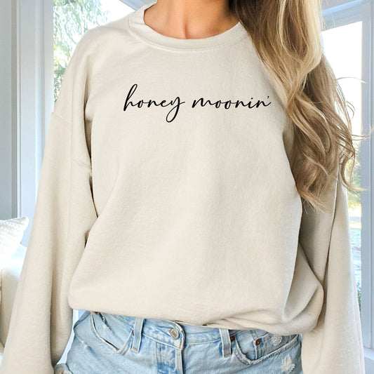 woman wearing a sand crewneck sweatshirt with honey moonin' printed across the chest in a black script font