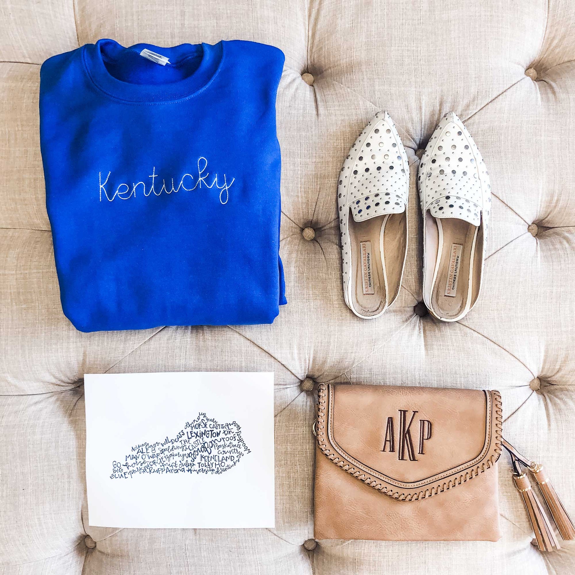 flatlay of outfit including a crossbody purse, white slides, and a royal blue crewneck with custom state stitched embroidery in white thread across the chest.