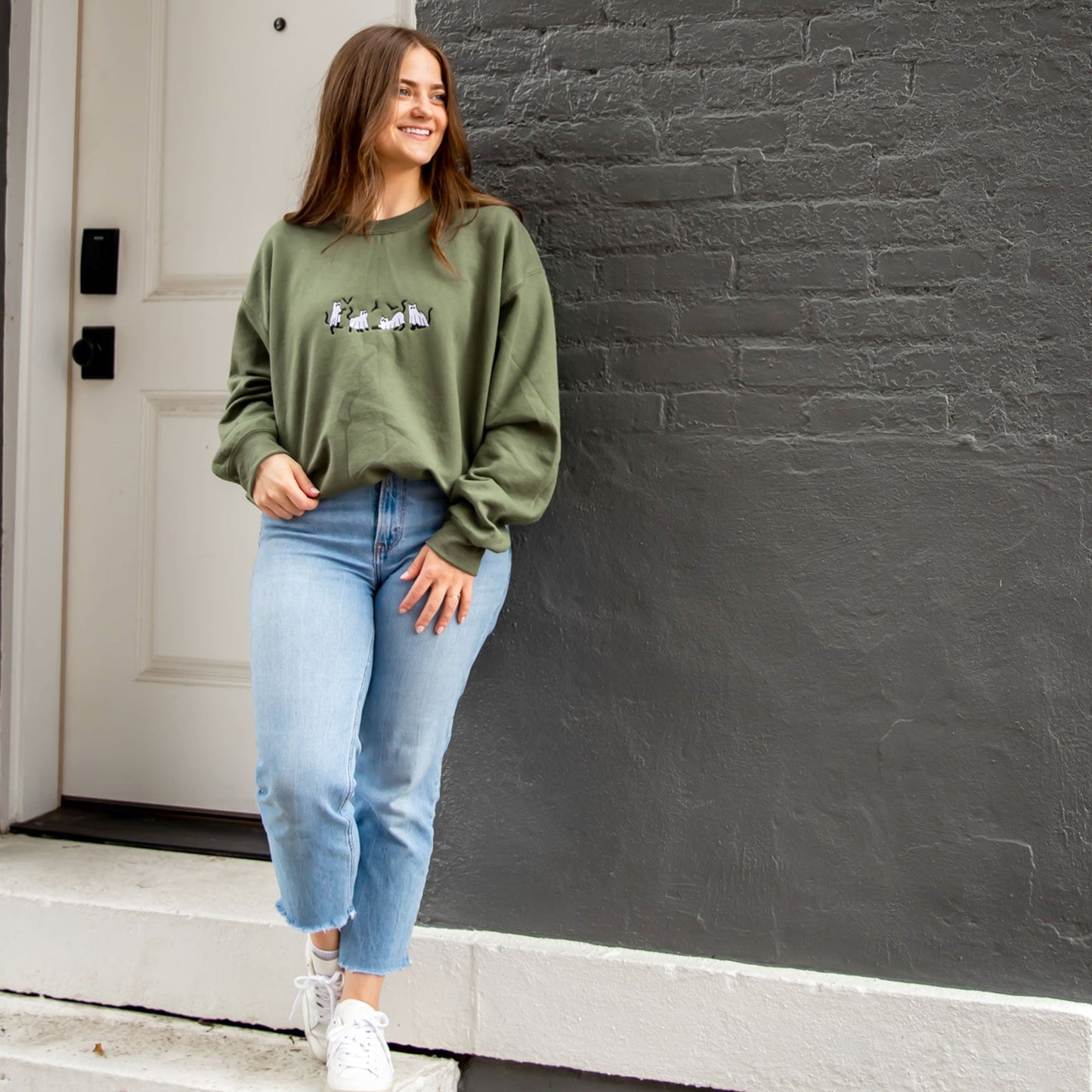 young woman wearing a military green crewneck sweatshirt with embroidered ghost cats design across the chest