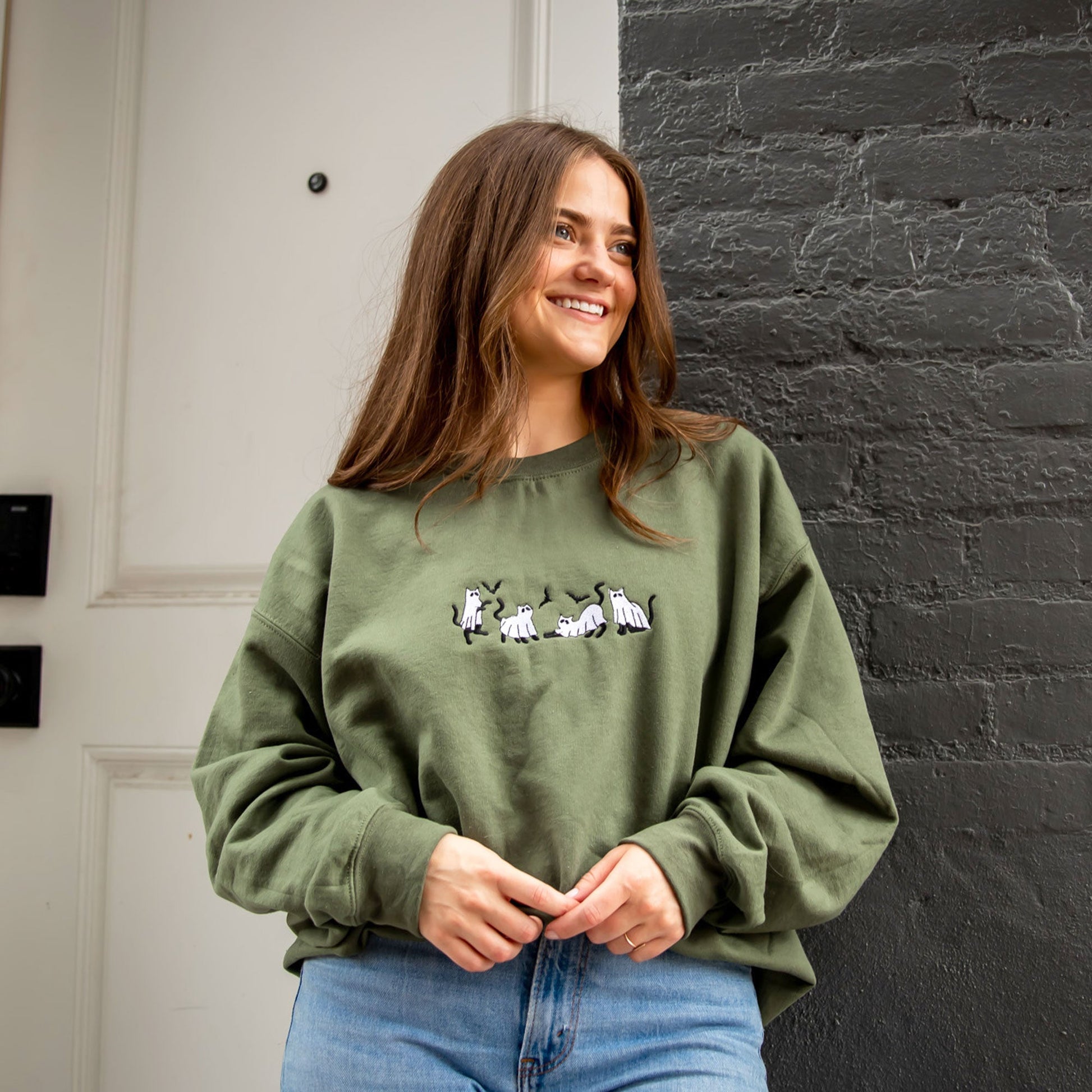 young woman wearing a military green crewneck sweatshirt with embroidered ghost cats design across the chest