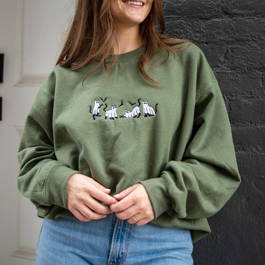 young woman wearing a military green crewneck sweatshirt with embroidered ghost cats design across the chest