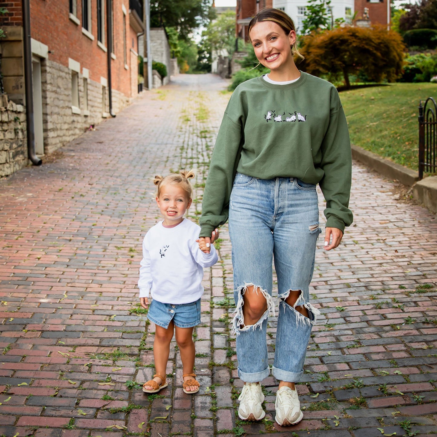 young woman wearing a military green crewneck sweatshirt with embroidered ghost cats design across the chest with her young daughter in a white crewneck sweatshirt with embroidered ghost cat