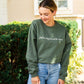 woman wearing jeans and a military green crewneck sweatshirt with a custom embroidery reading 'fall is my favorite color' in a natural thread color and a script font, standing outside a large cream brick house.