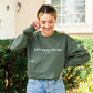 woman wearing jeans and a military green crewneck sweatshirt with a custom embroidery reading 'fall is my favorite color' in a natural thread color, standing outside a large cream brick house.