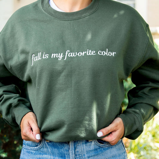 woman wearing jeans and a military green crewneck sweatshirt with a custom embroidery reading 'fall is my favorite color' in a natural thread color, standing outside a large cream brick house.