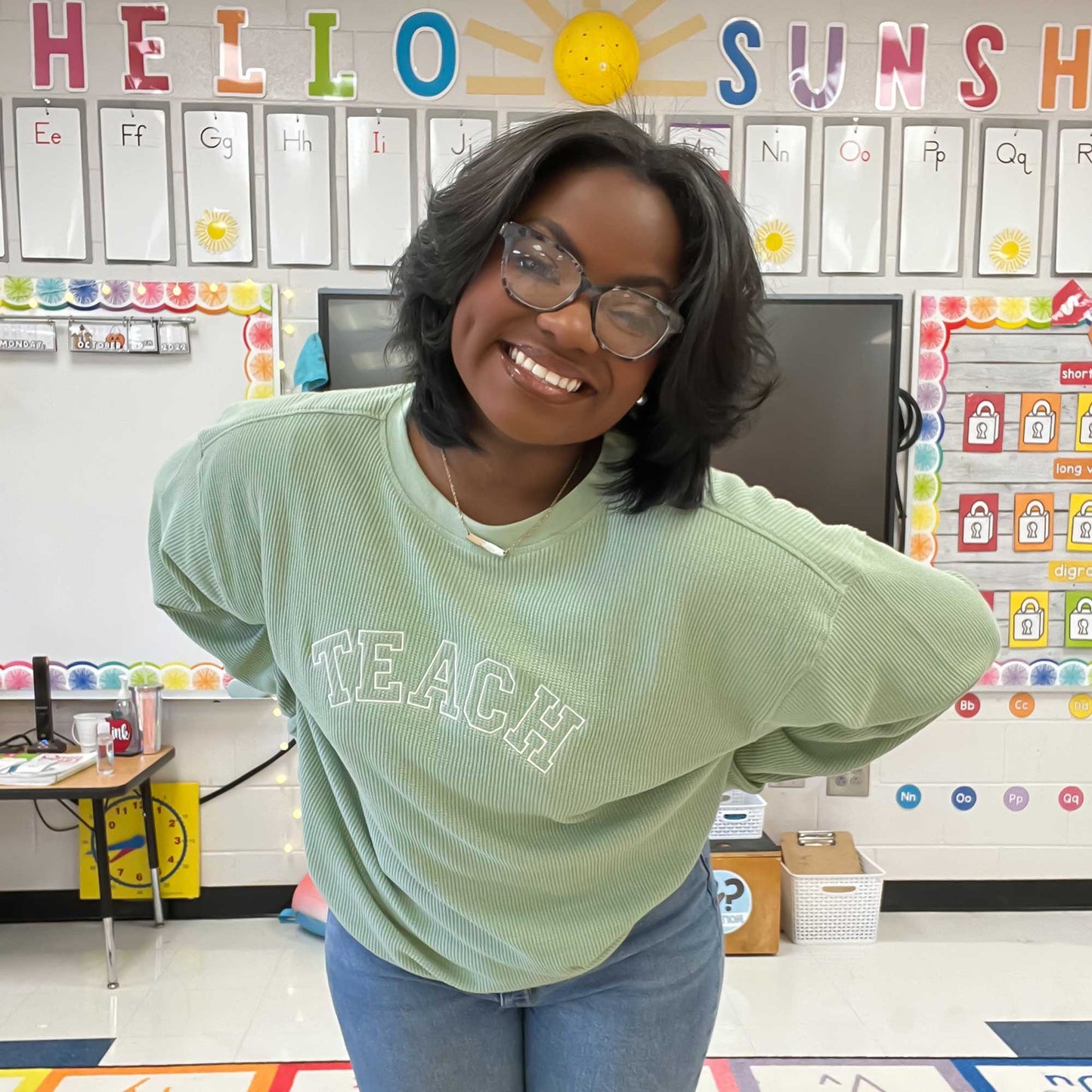 teacher in her classroom wearing a sage waffle knit crew with embroidered TEACH design