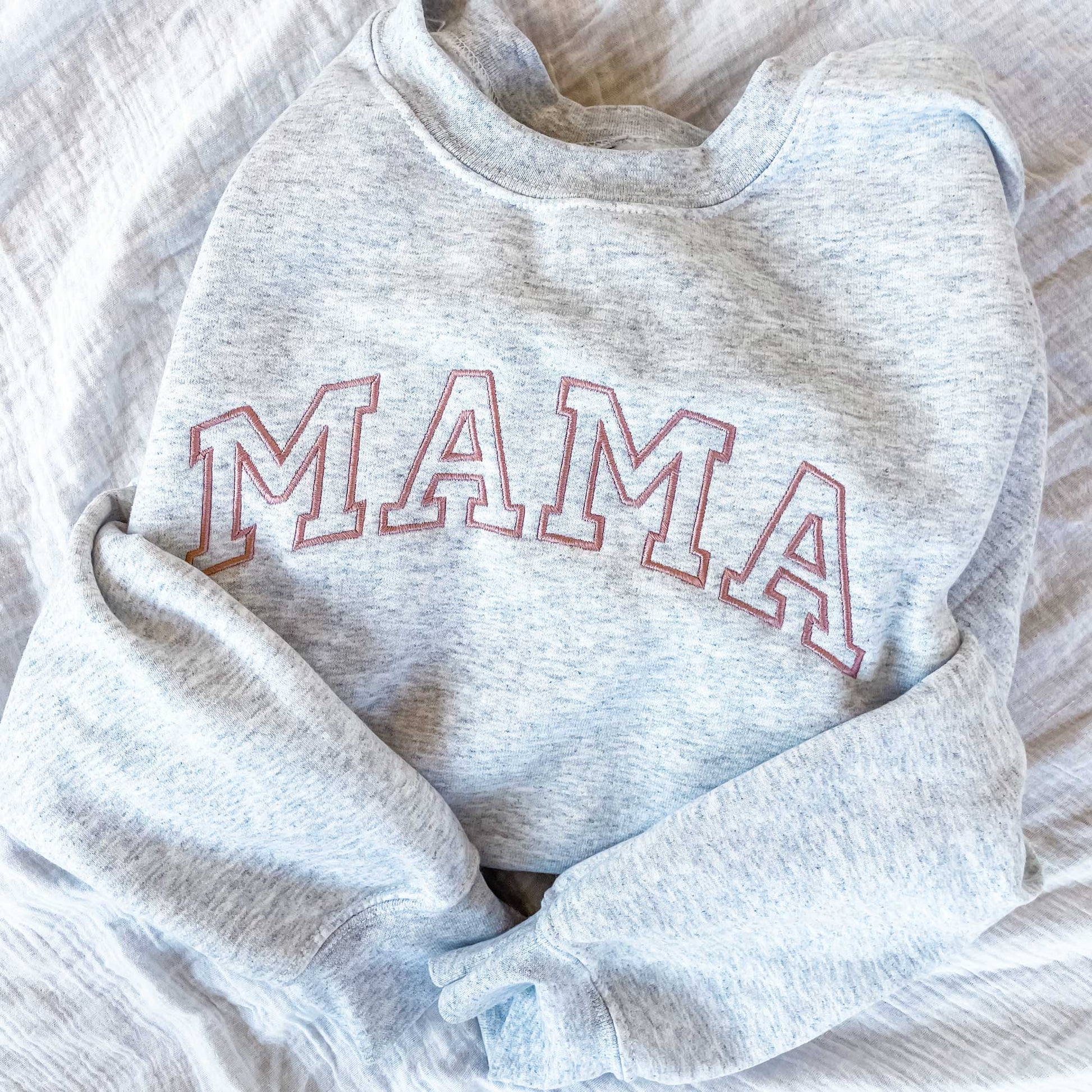 a light gray sweatshirt folded and laying on a bed featuring mama embroidered across the chest in a dark pink thread