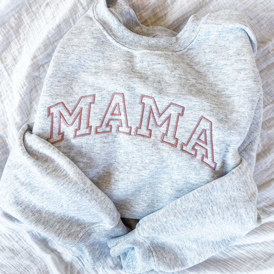 a light gray sweatshirt folded and laying on a bed featuring mama embroidered across the chest in a dark pink thread