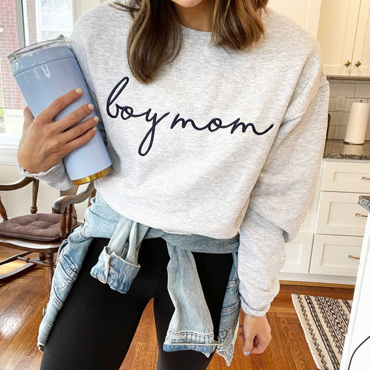 a mom standing in her kitchen wearing leggings, a jean jacket tied around her waist, and an ash gray crewneck sweatshirt with boy mom embroidered in a large script across the chest