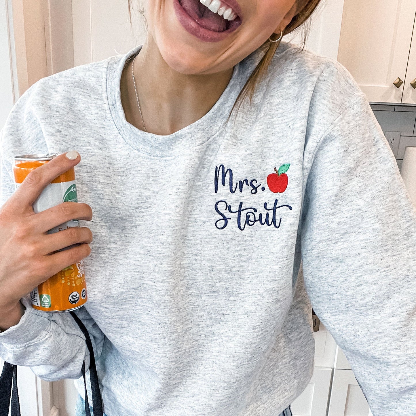 woman smiling and wearing a sweatshirt with a teacher's name and small apple embroidered on left chest
