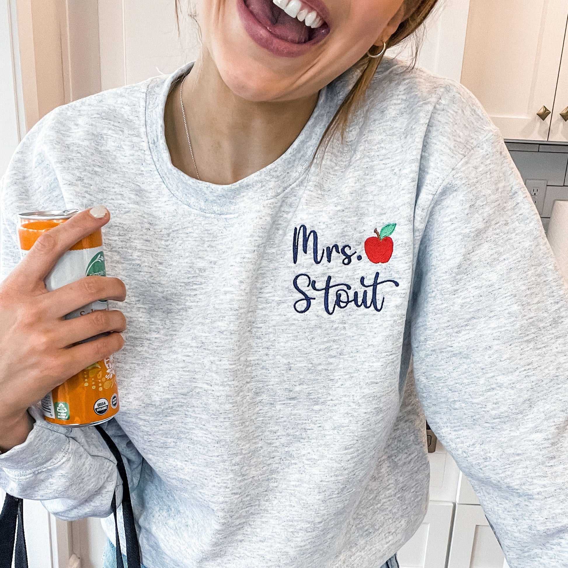 woman smiling and wearing a sweatshirt with a teacher's name and small apple embroidered on left chest
