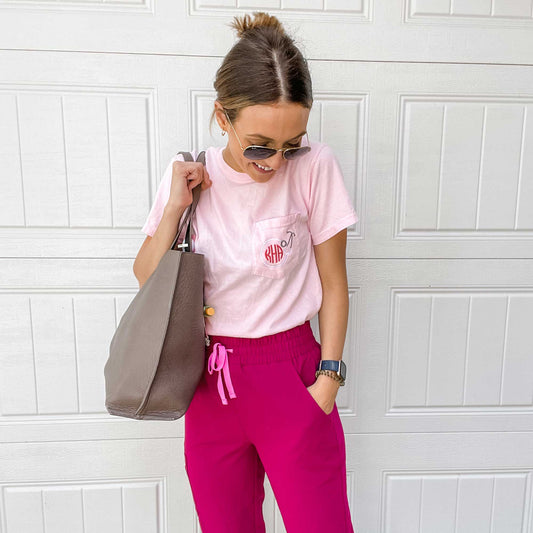 nurse holding a large tote bag and wearing hot pink scrub pants and a light pink t-shirt with a personalized embroidered design of a round stethoscope and monogram on the pocket