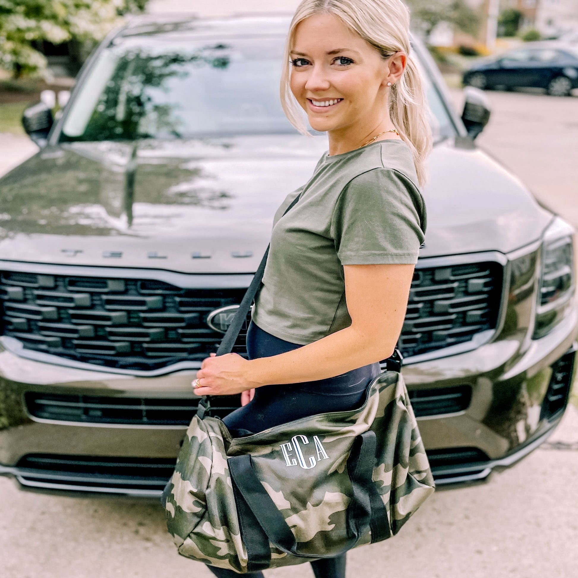 woman standing in front of car holding camo bag across chest