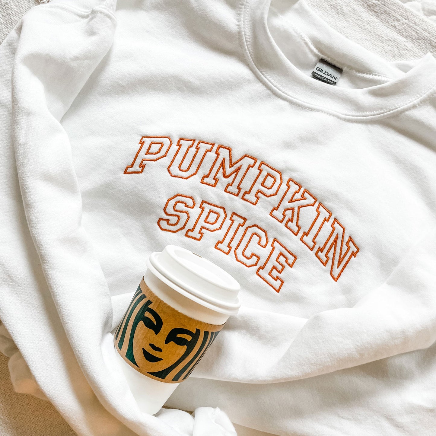 white sweatshirt folded and laying on top of a blanket with pumpkin spice embroidered on the front in athletic block style font