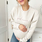 pregnant woman wearing a light beige crewneck sweatshirt with girl mama embroidered in a mauve thread across the chest
