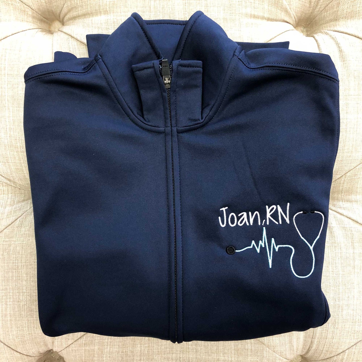 close up of a folded navy blue polyester full zip nurse jacket featuring a personalized name, credentials, and stethoscope embroidery 
