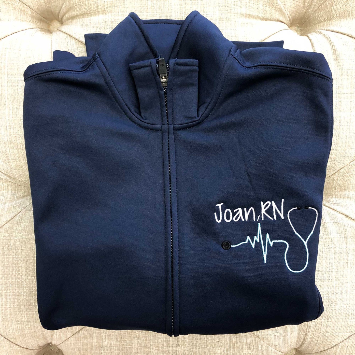 close up of a folded navy blue polyester full zip nurse jacket featuring a personalized name, credentials, and stethoscope embroidery