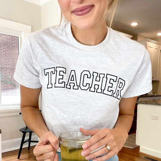woman in her home wearing a light gray crewneck top with teacher embroidered in block letters across the chest