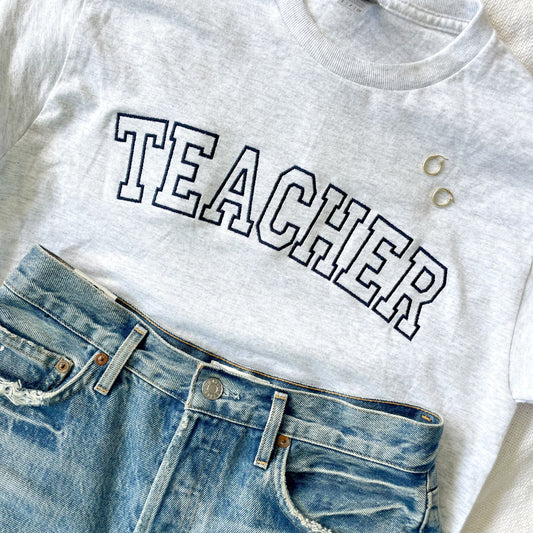 flat lay of ash crewneck tee with teacher embroidered in vintage athletic block letters across the chest