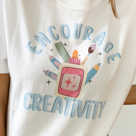 close up of a dtg printed design on a white t-shirt. the design has the word 'encourage'' arched over a grouping of colorful school supplies with 'creativitiy' underneath