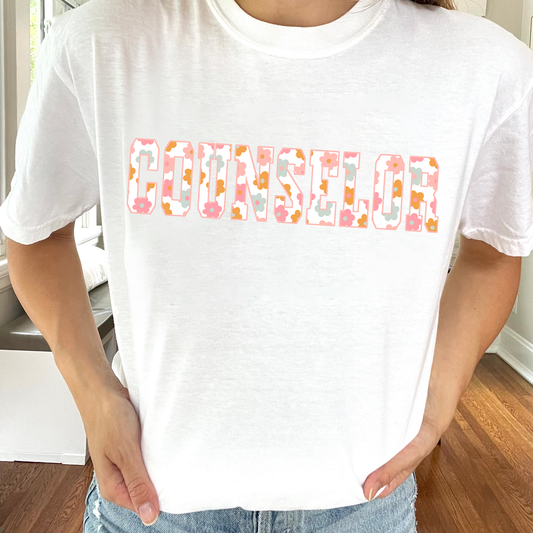 young woman wearing a white comfort colors t-shirt featuring a varsity COUNSELOR print with a pastel floral pattern fill