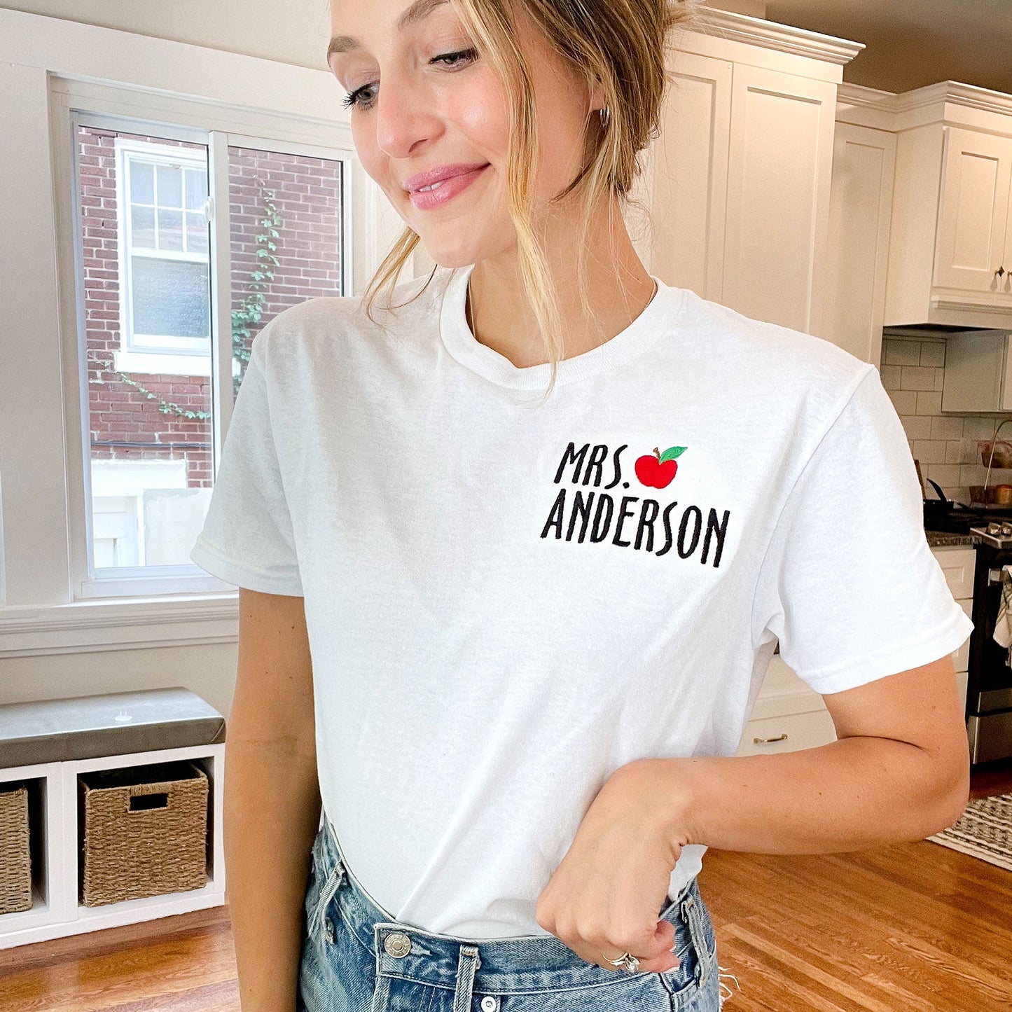 woman in her house looking down wearing white shirt with teacher mini apple embroidery