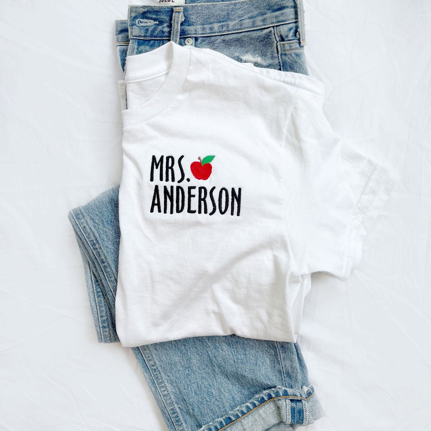 flat lay of mini apple teacher shirt on top of pair of jeans
