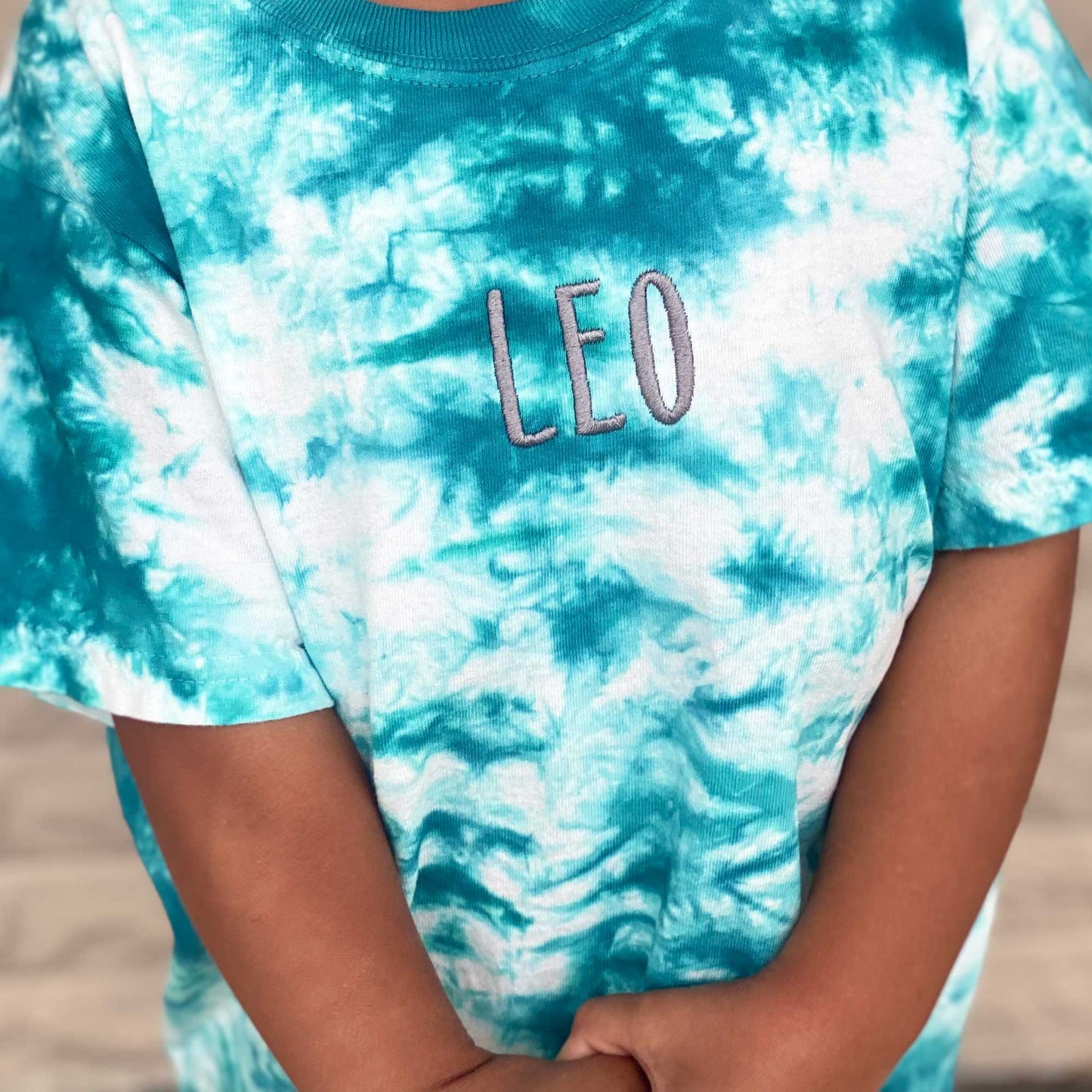 close up photo of a youth crystal tie dye t-shirt in a teal blue and white with a name embroidery in a dark gray thread on the center of the chest