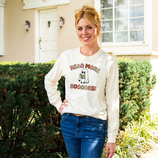 woman wearing a long sleeve ivory comfort colors tee with a dtg printed design. design reading 'read more boooks!' in a fall color pallet and a ghost reading a book