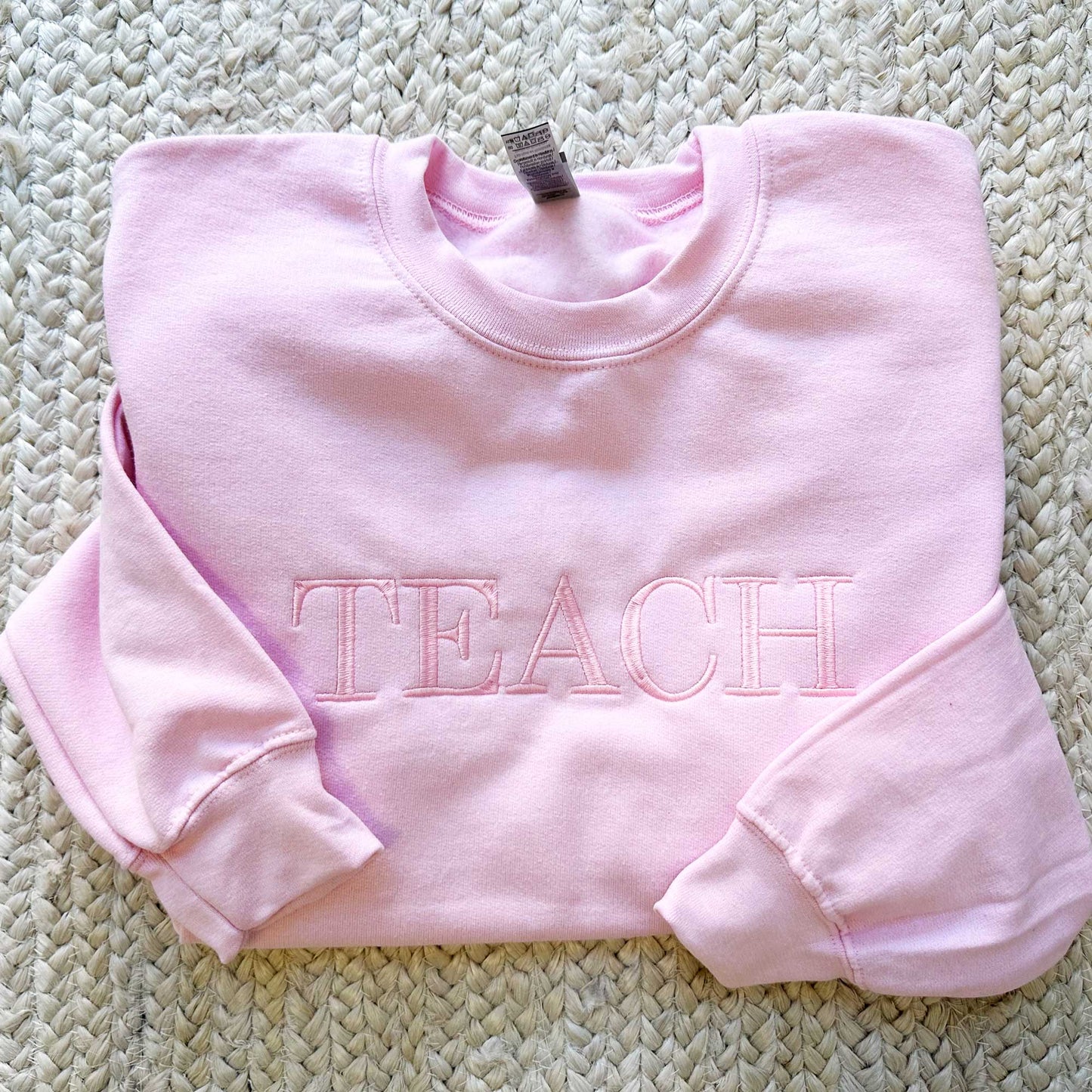 flat lay of an light pink crewneck sweatshirt with embroidered all caps teach in baby pink tone on tone thread