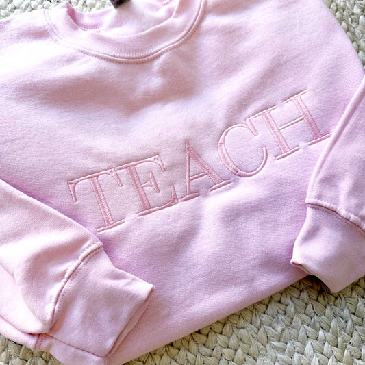 flat lay of an light pink crewneck sweatshirt with embroidered all caps teach in baby pink tone on tone thread