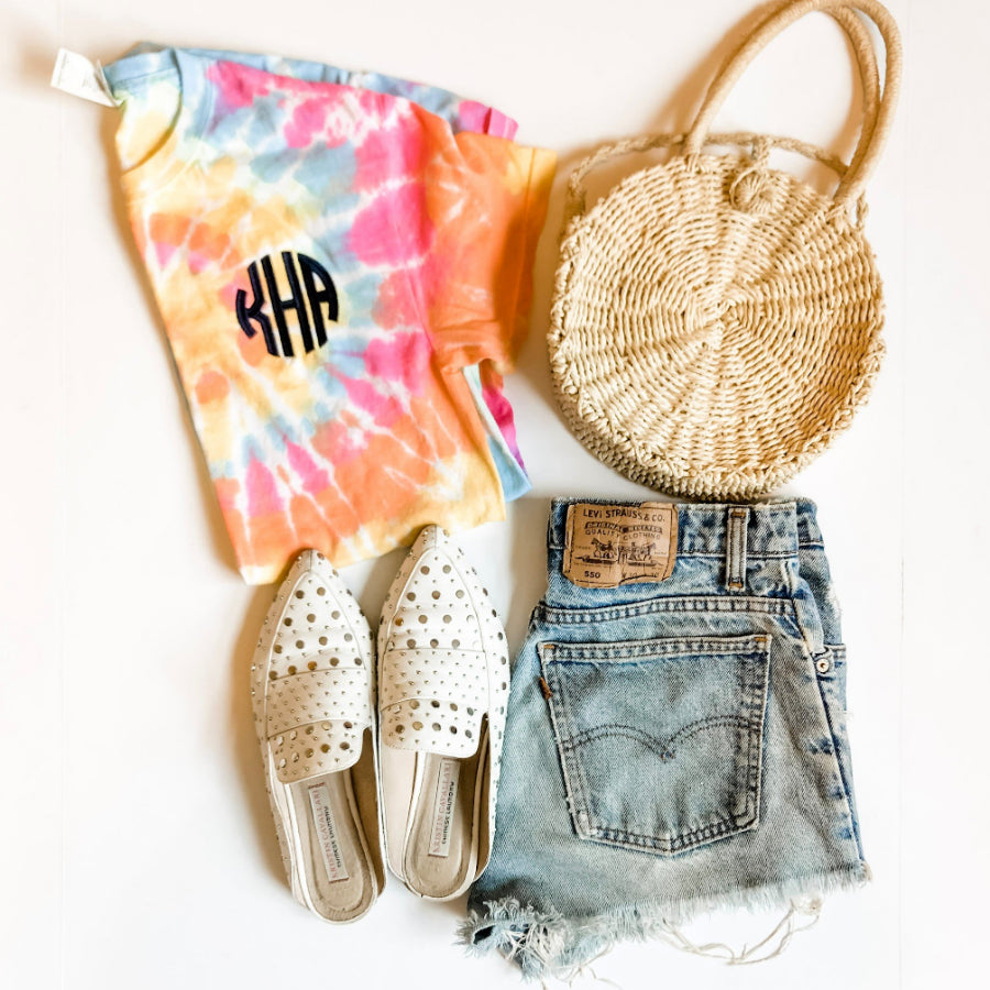 outfit flatlay with straw purse, jean shorts, sandals, and a monogrammed tie dye t-shirt